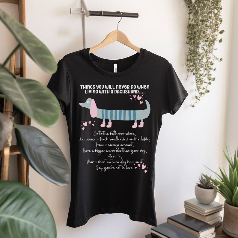 May include: Black t-shirt with a graphic of a dachshund wearing a striped shirt and the text "THINGS YOU WILL NEVER DO WHEN LIVING WITH A DACHSHUND.... Go to the bathroom alone, Leave a sandwich unattended on the table, Have a savings account, Have a bigger wardrobe than your dog, Sleep in, Wear a shirt with no dog hair on it, Say you're not in love."
