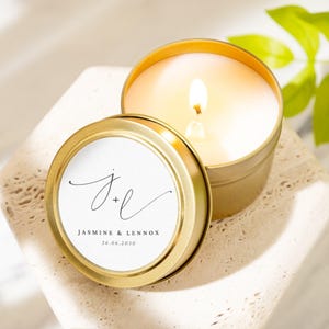 May include: A lit gold-colored candle in a round tin with a lid. The lid has a white label with the names "Jasmine & Lennox" and the date "26.06.2030" in black script. The candle sits on a textured, light-colored surface.