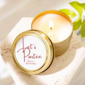 May include: A lit candle in a gold tin with a white label that reads "Let's Partee" in red script, with "Sarah's 40th birthday" below. The candle is on a textured, light-colored surface.