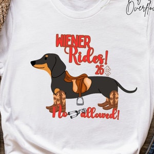 Op de afbeelding: Wit T-shirt met een zwarte teckel die cowboylaarzen en een zadel draagt. De tekst op het shirt luidt "Wiener Rides! 25¢ No Horses Allowed!"