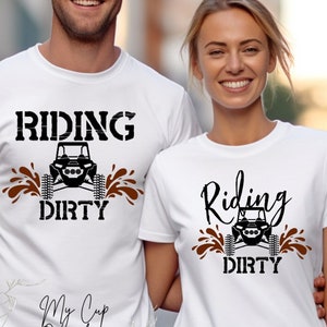 May include: Two white t-shirts with a black and brown graphic design. The graphic features a cartoon off-road vehicle with the text "Riding Dirty". One shirt has the text "My Cup Overflows" below the graphic.