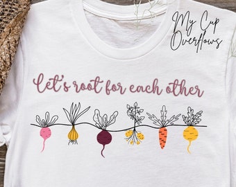 Könnte beinhalten: Weißes T-Shirt mit dem Schriftzug "Let's root for each other" in Schreibschrift. Darunter eine Reihe von illustrierten Wurzelgemüsen in Rosa, Gelb, Lila und Orange. Das Shirt wird mit einer blauen Jeans und einem braunen Hut kombiniert.