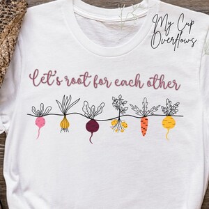 May include: White t-shirt with the text "Let's root for each other" in a cursive font. Below the text is a line of illustrated root vegetables in pink, yellow, purple, and orange. The shirt is styled with a pair of blue jeans and a brown hat.