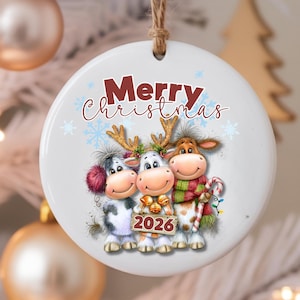 Può includere: Un ornamento rotondo in ceramica bianca appeso a uno spago di juta. Il design raffigura tre mucche dei cartoni animati con corna, accessori invernali e un cartello con la scritta 2026. Sopra di esse, il testo Merry Christmas in rosso e marrone.