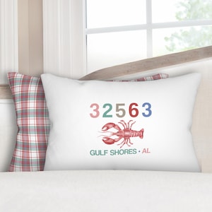 May include: White decorative pillow featuring a red lobster illustration and the text "32563 GULF SHORES • AL" in various colors. The pillow is placed in front of a window and a plaid pillow.
