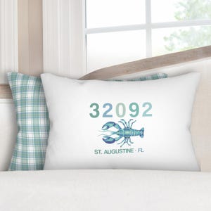 May include: White decorative pillow with the number "32092" in teal and a teal and blue lobster illustration. The text "ST. AUGUSTINE • FL" is below the lobster. A plaid pillow is visible in the background.