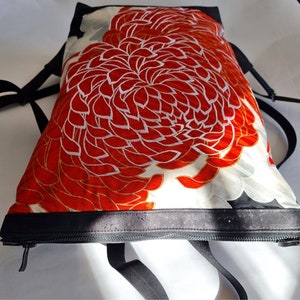 May include: A backpack with a vibrant floral design. The main fabric features a large red and white flower pattern with gold accents. The bag has black straps and a black zippered closure.