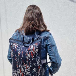 May include: A person wearing a blue denim jacket with a floral patterned backpack. The backpack has a dark blue background with white and red flowers.