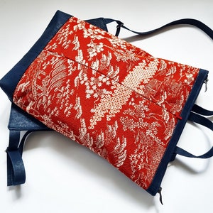 Puede incluir: Una bolsa de mano roja y blanca con estampado, con una correa y una cremallera azul marino. La bolsa tiene un diseño floral con flores y hojas blancas sobre un fondo rojo.