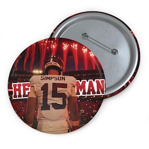 May include: A round button featuring a football player in a white jersey with the number 15 and the name "SIMPSON". The background shows a stadium with red fireworks and the text "HE MAN". The button has a silver pin back.