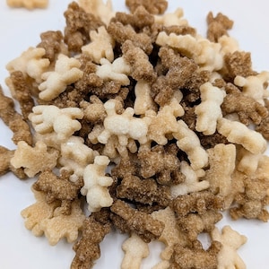 May include: A pile of bear-shaped cereal pieces in shades of light tan and brown. The cereal pieces are scattered on a white surface, with a few pieces isolated in the background. The shapes are detailed and textured.