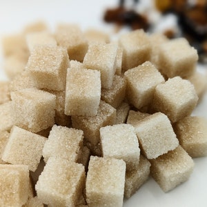 May include: A close-up shot of a pile of brown sugar cubes. The cubes are stacked together, with a few scattered around the edges. The sugar cubes have a slightly rough texture and a light brown color.
