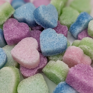 May include: A collection of heart-shaped sugar crystals in pastel shades of blue, green, and pink. The sugar crystals are scattered on a white surface.