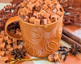May include: A glossy orange ceramic mug with embossed pumpkin designs is filled to the brim with small, light brown, cube-shaped sugar pieces. A gold-toned metal spoon with a leaf-shaped handle rests in front of the mug alongside star anise.