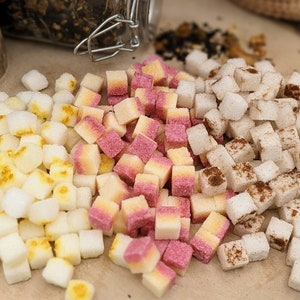 May include: An assortment of colorful, square-shaped candies arranged on a wooden surface. The candies are white, yellow, pink, and some are dusted with cinnamon. The image suggests a variety of sweet treats, possibly for a dessert or snack.