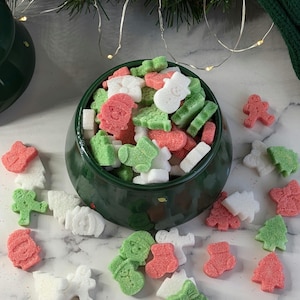 May include: A green bowl filled with white, green, and red sugar cubes shaped like Christmas trees, snowmen, and Santa Claus. The bowl is surrounded by more sugar cubes on a white marble surface.