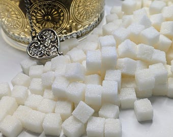 May include: Close-up of white sugar cubes scattered around a decorative gold and silver sugar bowl. The sugar cubes are small, square, and piled together. The sugar bowl has a heart-shaped charm.