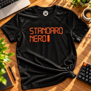 Peut inclure: Un t-shirt noir à manches courtes et col rond avec le texte orange "STANDARO NERO" imprimé sur la poitrine. Le t-shirt présente un petit logo circulaire sur l'étiquette intérieure du col représentant une empreinte de patte avec le texte "PAW PRINT".