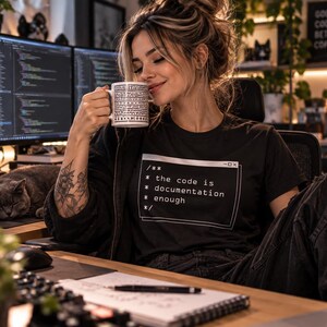 May include: A black t-shirt featuring a white graphic print of a code comment block that reads: /** * the code is * documentation * enough */. The shirt has a crew neckline and short sleeves, worn in a workspace setting with computer monitors.