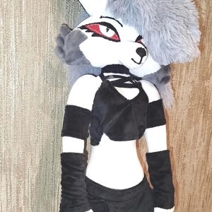 May include: A plush doll of a cartoon character with a white face, ears, and paws. The doll has a gray fur hairstyle, red eyes, and is wearing a black crop top, shorts, and arm warmers. The doll is approximately 20 inches tall.