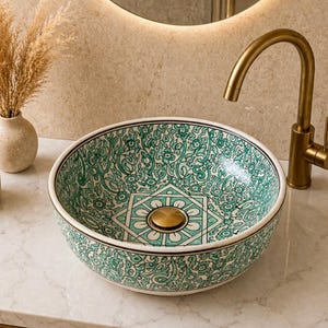 Könnte beinhalten: Ein rundes Aufsatzwaschbecken aus Keramik mit einem detaillierten grün-weißen Blumenmuster und einem geometrischen Design in der Mitte. Das Waschbecken hat einen goldenen Ablaufstopfen und eine passende goldene Armatur.
