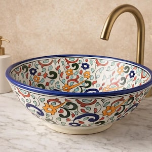 Peut inclure: Un lavabo en céramique décoratif avec un motif floral vibrant en orange, bleu et vert sur fond blanc. Le bord est souligné de bleu. Un robinet et un distributeur de savon dorés sont visibles.