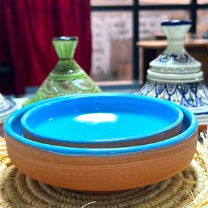 Può includere: Una pila di ciotole in terracotta con interno e bordo turchese vibrante, su un tappetino di paglia intrecciata. Pezzi di ceramica decorativi in verde e bianco, e blu e bianco sullo sfondo.