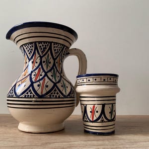 Peut inclure: Une cruche en céramique blanche et une tasse assortie avec des motifs géométriques bleus, verts et orange. La cruche a une anse et une large ouverture. La tasse est plus courte et a une forme cylindrique.