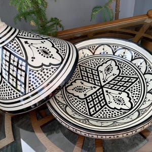 Peut inclure: Une tagine en céramique blanche avec un motif géométrique noir et blanc. La tagine a une base ronde et un couvercle conique. Le couvercle a un petit bouton sur le dessus.