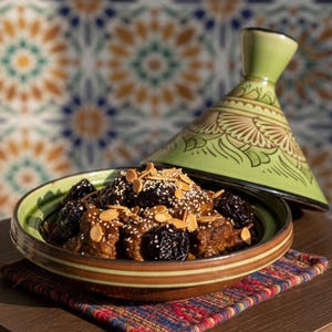 May include: A traditional Moroccan tagine dish with a green and brown ceramic lid. The dish contains meat, prunes, and almonds, served in a brown and green bowl. The background features a colorful mosaic tile pattern.