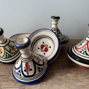 Peut inclure: Quatre tagines marocains en céramique avec des motifs floraux et géométriques colorés. Les tagines sont blancs avec des accents bleus, rouges, verts et noirs.