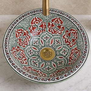 Peut inclure: Un évier décoratif en céramique avec un motif floral rouge, vert et blanc détaillé. L'évier a un drain et un robinet dorés. Un distributeur de savon et un vase blancs sont visibles en arrière-plan.