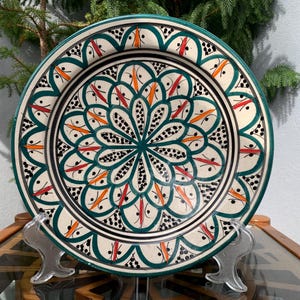 Peut inclure: Une grande assiette ronde en céramique avec un fond blanc et un motif géométrique vert, orange et noir. L'assiette est sur un support.