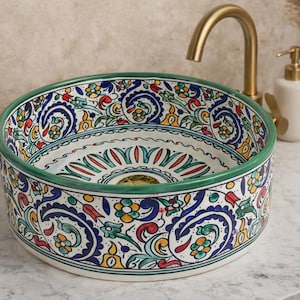 Peut inclure: Un évier rond en céramique avec un motif floral vibrant en bleu, rouge, jaune et vert. L'évier a un bord vert et est placé sur un fond neutre. Un robinet doré et un distributeur de savon blanc sont visibles.