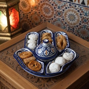 May include: A decorative serving tray with a blue and white patterned design, filled with various pastries and a central lidded dish. The tray sits on a wooden table, with a colorful lantern and mosaic tile background.