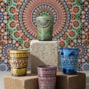 Peut inclure: Quatre verres à boire en céramique peints à la main, aux couleurs vives. Les verres sont jaunes, verts, bleus et roses, chacun avec des motifs uniques. Ils sont présentés sur une surface neutre devant un fond de mosaïque.