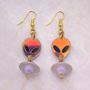 May include: A pair of dangle earrings featuring a gold-tone hook, an iridescent alien head charm with black oval eyes, and a translucent, milky white circular bead at the base. The alien charms display shifting orange, pink, and yellow hues.