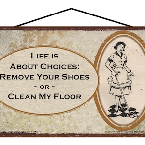 May include: A vintage-style sign with a distressed, beige background and brown text. The sign reads "Life is about choices: Remove your shoes or clean my floor." A cartoon image of a woman with a mop is in the corner of the sign.