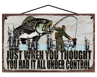 May include: A rustic wooden sign with a distressed white background featuring a cartoon illustration of a fisherman reeling in a large fish. The text on the sign reads "JUST WHEN YOU THOUGHT YOU HAD IT ALL UNDER CONTROL".