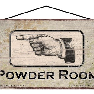 May include: A rectangular wooden sign with a distressed finish. It features a black and white illustration of a hand pointing to the right, above the words "POWDER ROOM". The sign includes the text "Hip Signs for Cool Folks" and "Made in USA".