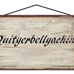 May include: Wooden sign with the phrase "Quityerbellyachin'" in black script. The sign has a distressed, vintage look with a cream-colored background and a dark brown border. A black string is attached for hanging.