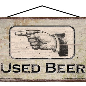 May include: A rustic wooden sign with a distressed, faded look. A hand points to the left with the text "USED BEER" in black lettering. The sign has the text "737L-58 Hip Signs for Cool Folks™" and "Lorrie Powell Studios Made in U.S.A" in smaller lettering.