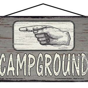 May include: A rustic wooden sign with a distressed gray finish. The sign features a white pointing hand graphic and the text "CAMPGROUND" in white lettering. The sign has a black hanging rope and two black metal hanging hooks.
