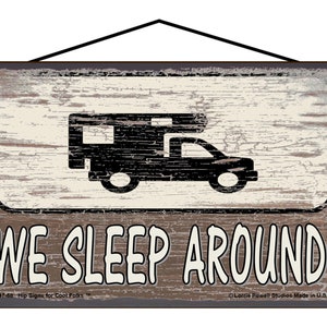 May include: A rustic wooden sign with a distressed white background and black text that reads "WE SLEEP AROUND". A black silhouette of a camper van is above the text. The sign has a distressed wood grain texture and a black rope for hanging.