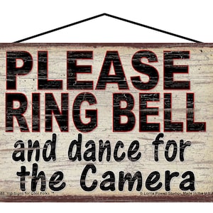 May include: A rustic wooden sign with distressed white paint and brown edges. The sign reads "PLEASE RING BELL and dance for the Camera". The text is in black with red outlines. The sign has a hanging hole at the top and the text "07X-58 Hip Signs for Cool Folks" is printed in the bottom left corner.