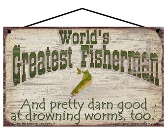 May include: A rustic wooden sign with a distressed white background and green text that reads "World's Greatest Fisherman And pretty darn good at drowning worms, too."  The sign features a cartoon image of a fish and has a rope for hanging.