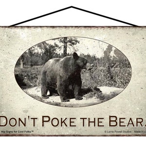May include: A vintage-style metal sign with a black and white photo of a bear in a forest setting. The sign reads "Don't Poke The Bear."  The sign is distressed and has a rustic look.