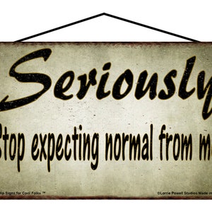 May include: A rectangular sign with the words "Seriously Stop expecting normal from me" in black script. The sign has a distressed, vintage look with a cream background and a black hanging cord.