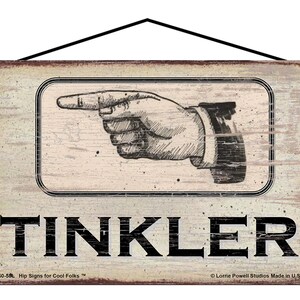 May include: A rustic wooden sign with a distressed white background and black text. A hand points to the right with the word "TINKLER" below. The sign has the text "Hip Signs for Cool Folks" and "Lorrie Powell Studios Made in U.S.A." at the bottom.