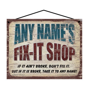 May include: A rectangular wooden sign with the text "ANY NAME'S FIX-IT SHOP" in blue and red lettering. The sign includes the phrases "IF IT AIN'T BROKE, DON'T FIX IT" and "BUT IF IT IS BROKE, TAKE IT TO ANY NAME!" The sign has a distressed, vintage look.